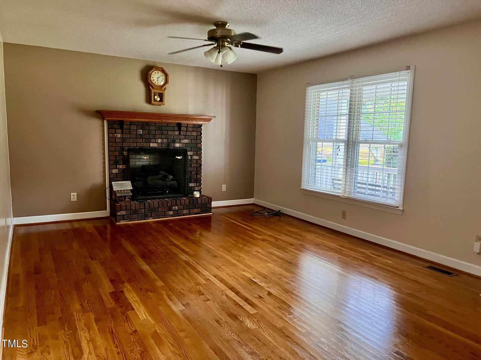 A cozy living room with polished wooden floors, a brick fireplace with a wooden mantel, a wall clock above, and a ceiling fan. Large window with blinds on the right lets in natural light. Walls are painted in neutral tones.