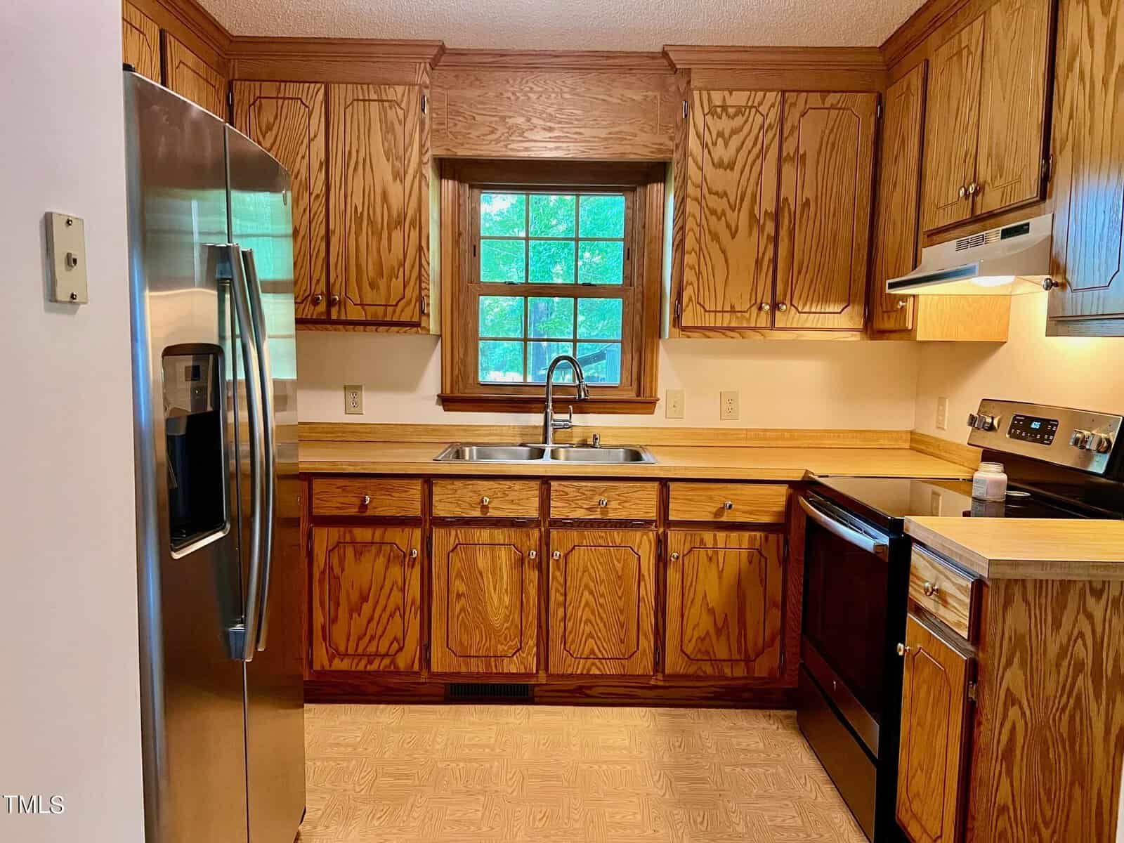 A kitchen with wooden cabinets and a matching counter. It features a stainless steel refrigerator, a stove with an oven, and a double sink situated under a window with a view of greenery outside.