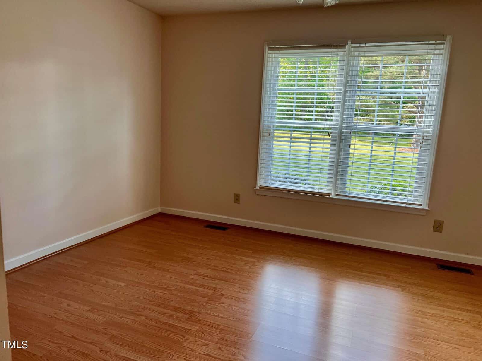 An empty room with light brown wooden floors, cream-colored walls, and a large window with white blinds. Sunlight filters through the window, illuminating the floor.