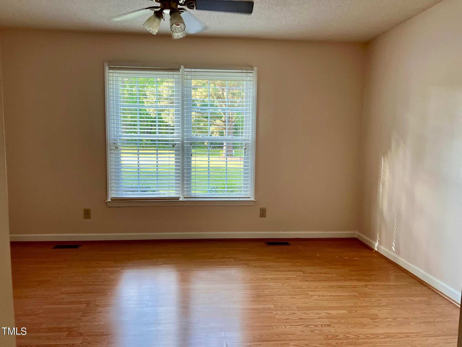 Empty room with light brown hardwood floors, beige walls, and a ceiling fan. Large window with white blinds letting in natural light, overlooking a grassy outdoor area.