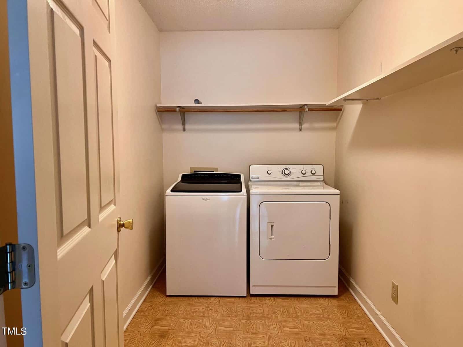 A laundry room with a washing machine and dryer side by side. The walls are beige, and there is a wooden shelf above the appliances. The floor has a light brown tile pattern, and an open door is on the left.