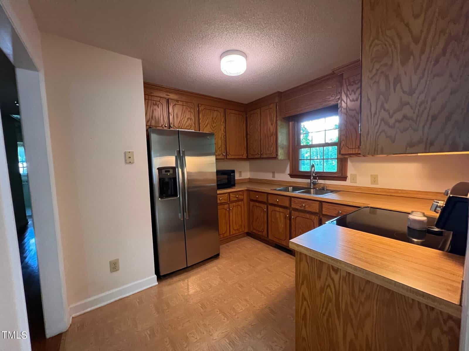 A kitchen with wooden cabinets, a stainless steel refrigerator, and a built-in microwave. The counter space is ample, and a window above the sink provides natural light. The floor has a light wood finish.