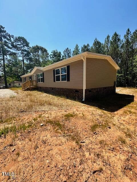A beige mobile home with a small front porch sits on a dry dirt patch surrounded by sparse vegetation and tall trees under a clear blue sky.
