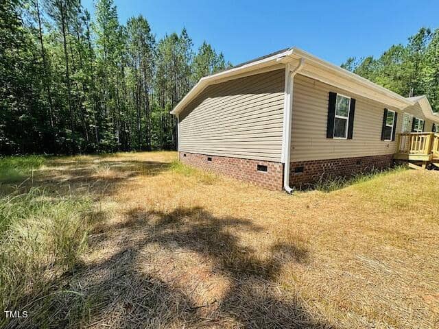 A beige house with a brick foundation is surrounded by dry grass and tall trees under a clear blue sky. A small wooden porch is visible on the right.