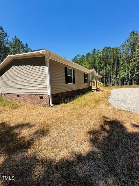 A single-story house with beige siding and a brick foundation sits in a grassy area. There's a small wooden porch with stairs leading to the entrance. A gravel driveway is visible, and tall trees surround the property under a clear blue sky.
