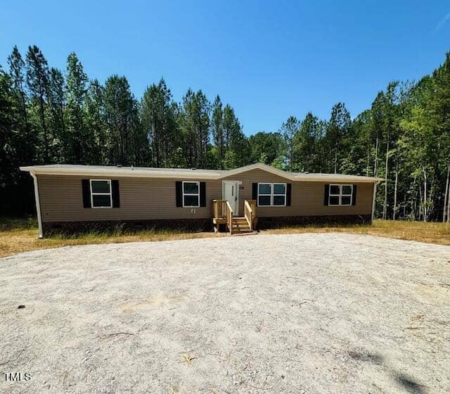 A beige single-story mobile home with white trim sits on a gravel driveway surrounded by tall trees and greenery. The home features a small wooden porch and several windows under a clear blue sky.
