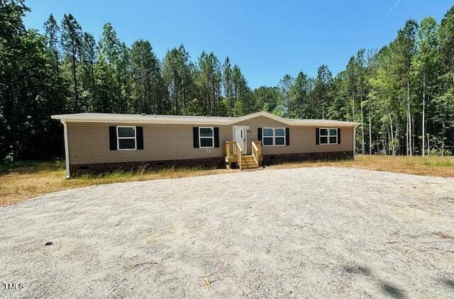 A single-story beige manufactured home sits in a clearing surrounded by tall trees. It has a simple front porch with steps and a gravel driveway leading up to it. The sky is clear and sunny.