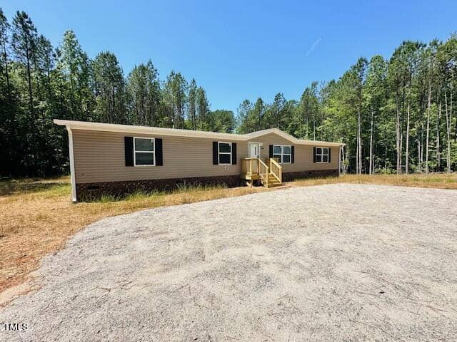 A single-story manufactured home with beige siding sits on a spacious, cleared plot at 4506 Nc Hwy 56 in Franklinton. Surrounded by tall trees under a clear blue sky, a small wooden deck leads to the front entrance.