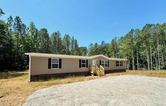 A single-story, beige mobile home with dark shutters and a small front porch sits on a grassy plot surrounded by tall trees under a clear blue sky. A gravel driveway leads up to the house.