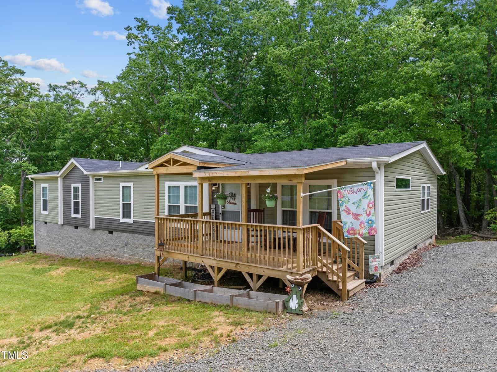 A ranch-style house with a mix of beige and green siding is surrounded by lush trees. It features a wooden porch with potted plants and a decorative quilt hung on the railing. A gravel driveway leads up to the entrance.