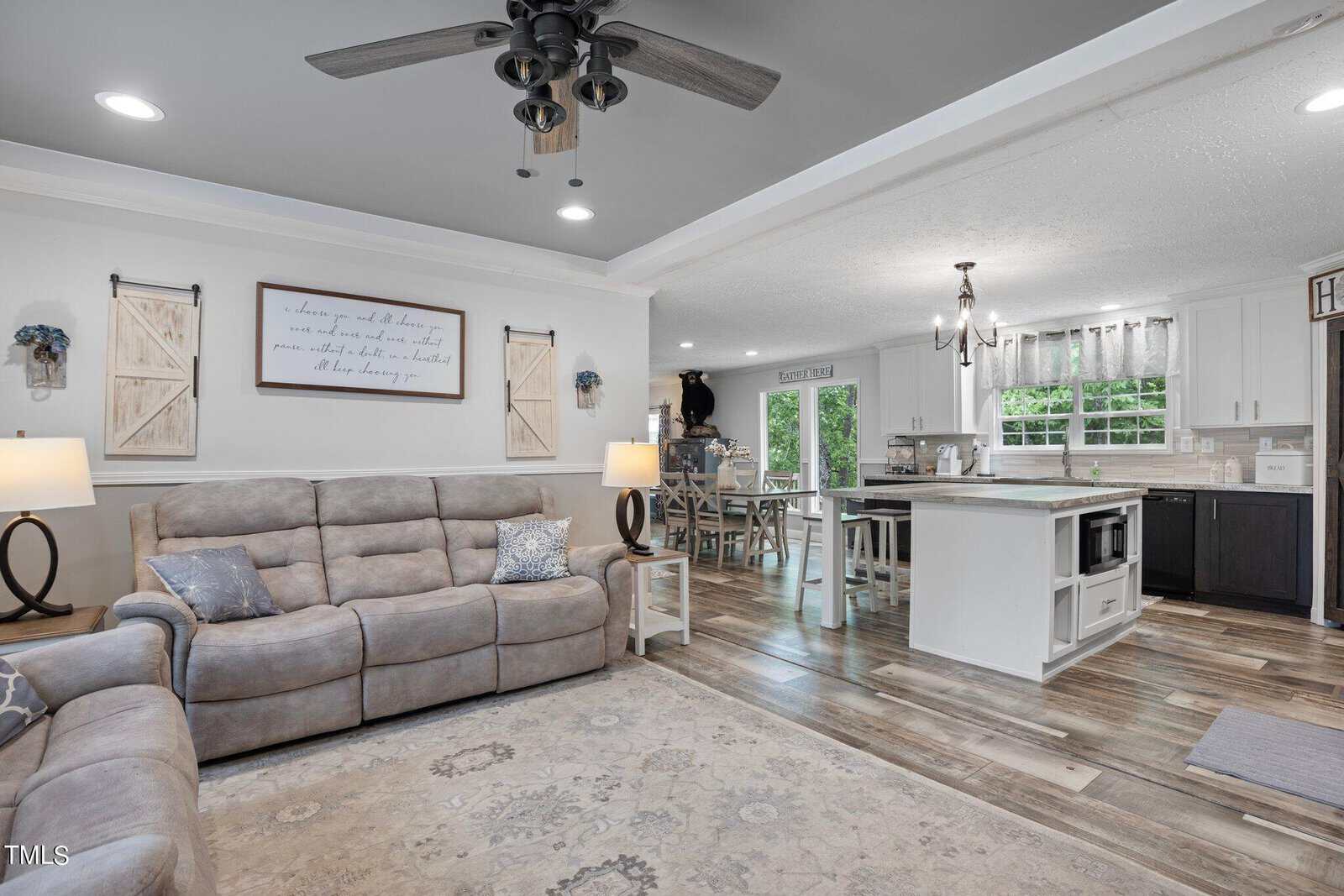 A cozy, open-plan living room and kitchen with rustic decor. The living area has a plush gray recliner sofa and a wooden coffee table. The kitchen features a large island, white cabinets, and a dining table. Wood flooring and light fixtures complete the space.