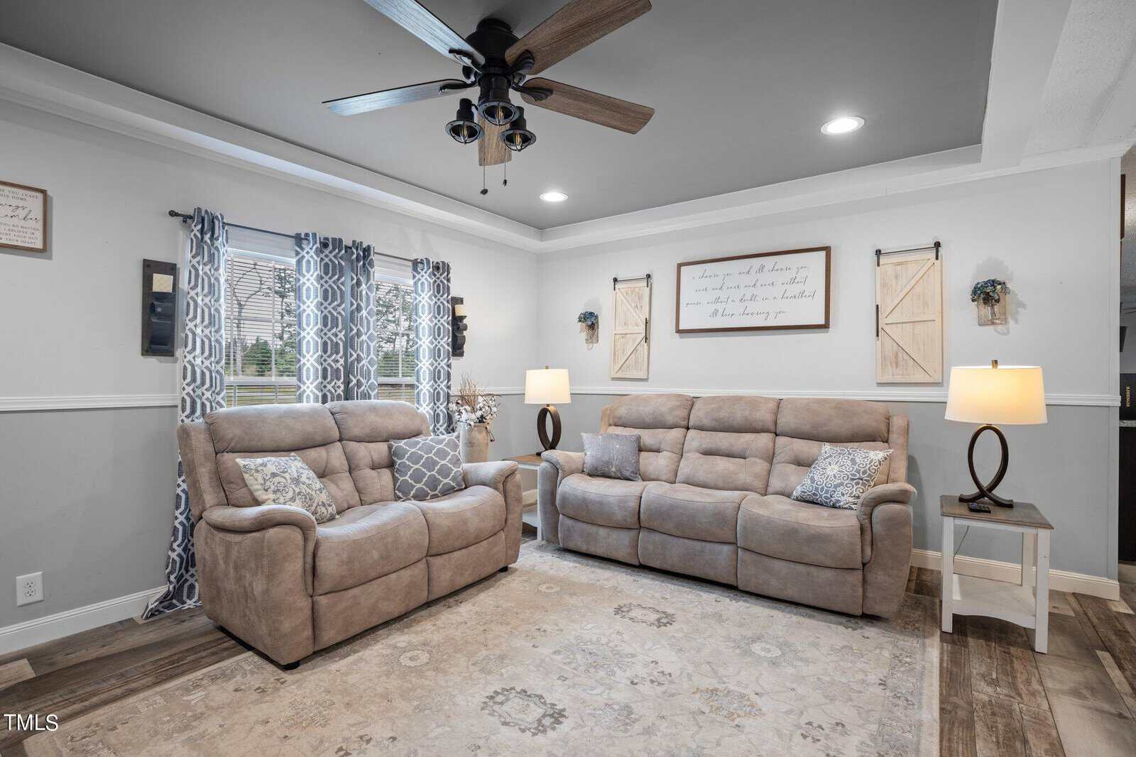 A cozy living room with beige reclining sofas, a patterned rug, and two table lamps. Decorative wall art and curtains adorn the room. A ceiling fan hangs above, and natural light filters through the window.
