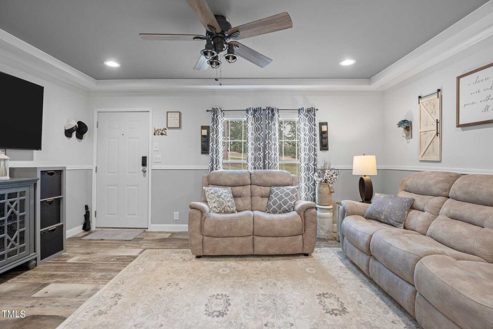 Living room with beige sofas, a large rug, and a ceiling fan. Curtains frame double windows. Decor includes wall art and a lamp. The door is on the left, and the flooring is light wood.