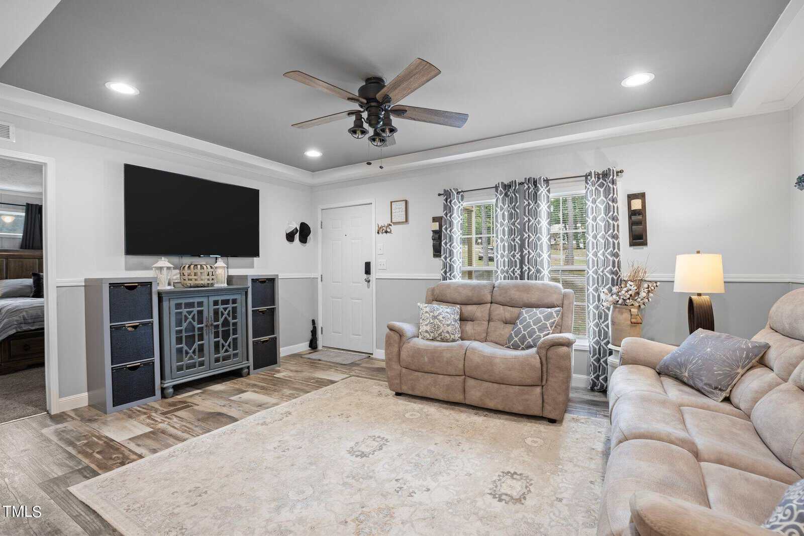 A cozy living room with a beige sectional sofa and matching loveseat. A large black TV is mounted above a dark wood console with decorative items. Gray curtains adorn windows, and a ceiling fan is overhead. A light patterned rug covers the floor.