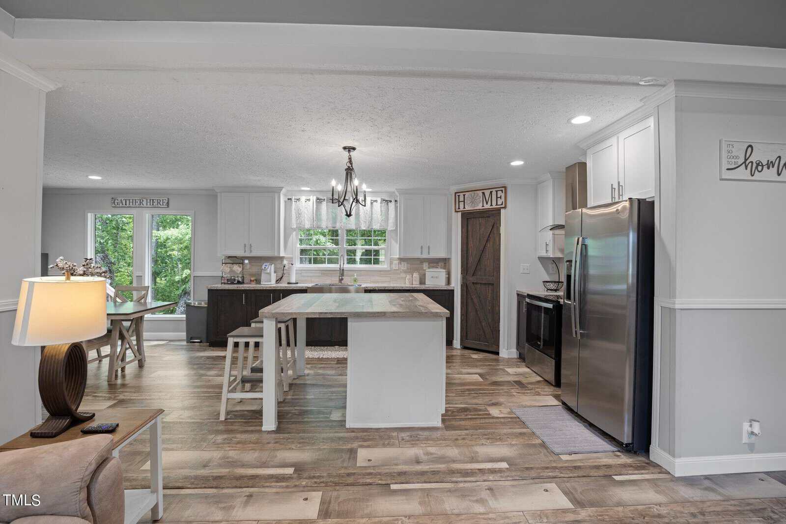 A modern kitchen with a central island, white cabinetry, and stainless steel appliances. A wooden dining table with chairs is visible near a large window. A rustic sliding barn door leads to an adjacent area. A chandelier hangs above the island.