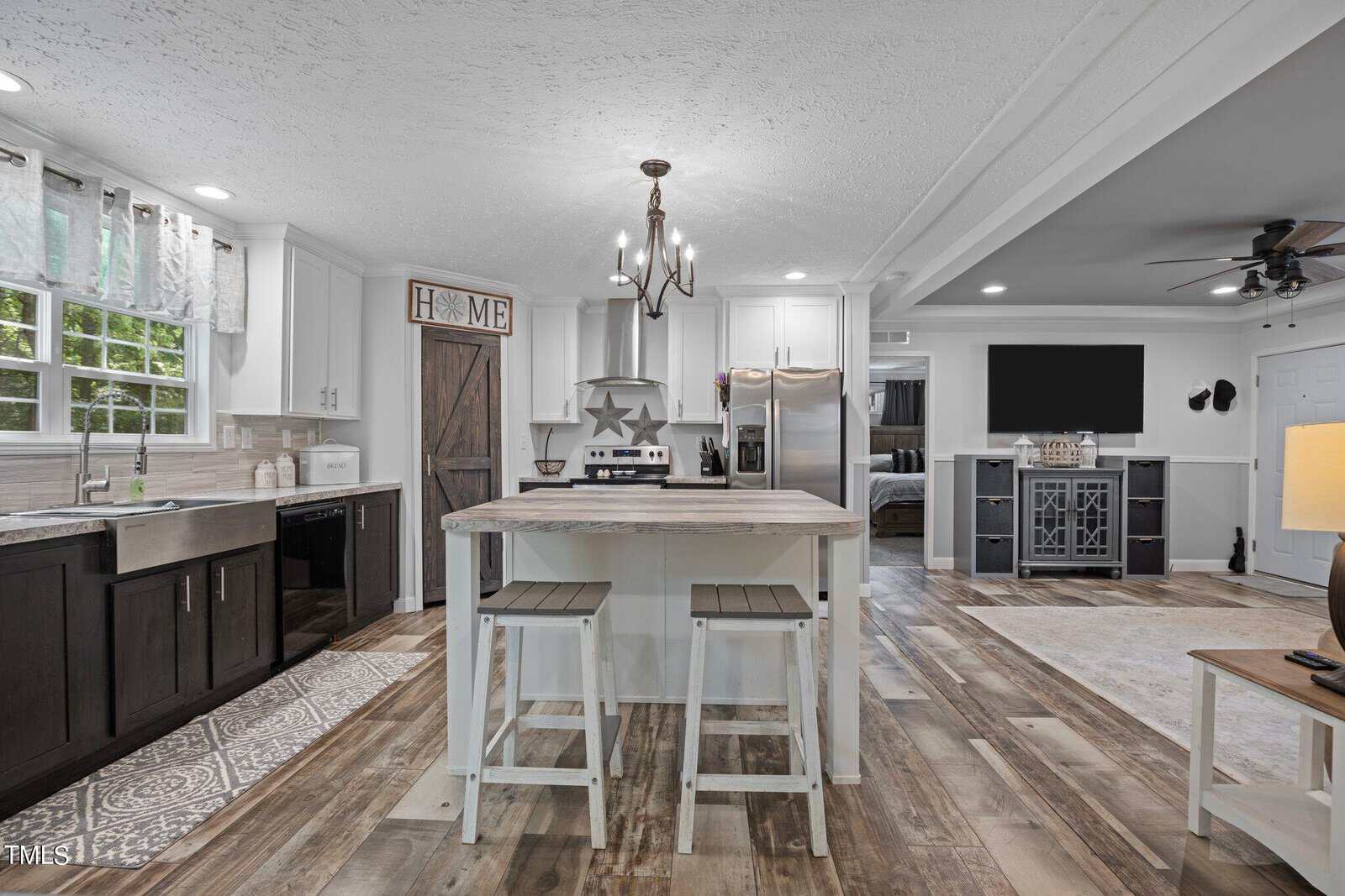 A modern kitchen with a central island featuring two stools. The space includes a stove, stainless steel refrigerator, and cabinets. A rustic door with "Home" decor hangs on the wall. Light wood flooring runs throughout the open-plan area.