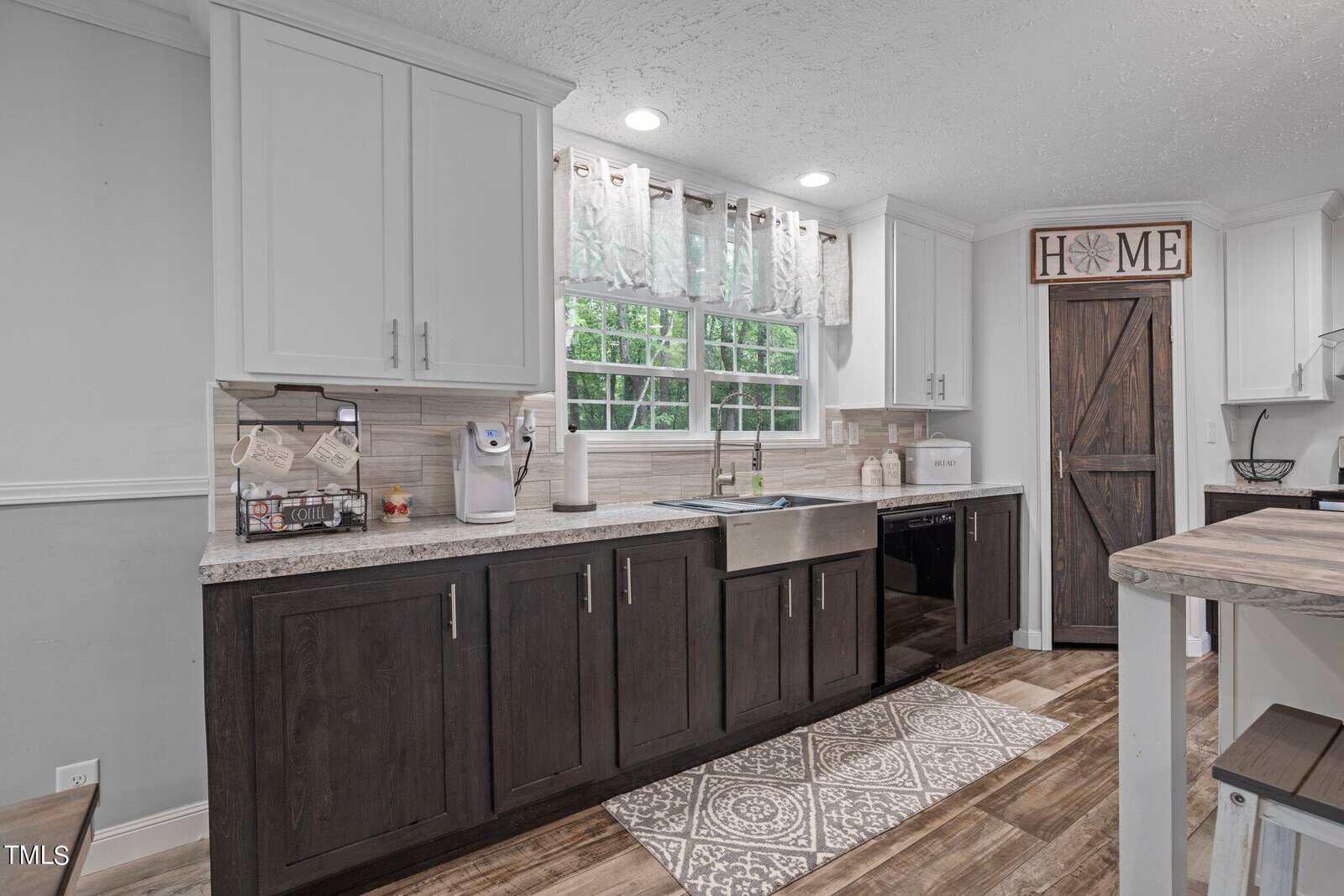 A modern kitchen with a farmhouse style features white upper cabinets and dark lower cabinets. A farmhouse sink is set beneath a large window with decorative curtains. The room includes rustic wooden accents and patterned floor mats.