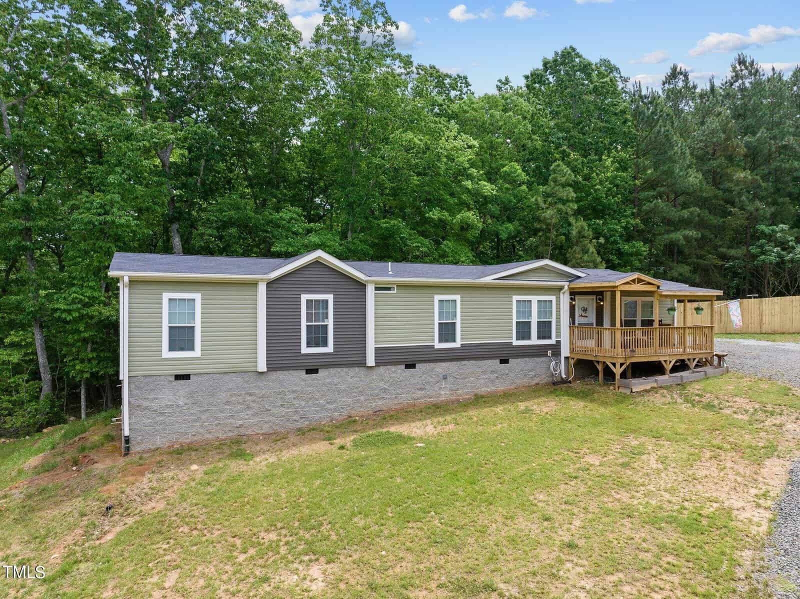 Nestled at 7074 Mountain Road in Oxford, this charming single-story manufactured home features gray and beige siding. It boasts a wooden porch on the right side, perfect for enjoying the view of dense green trees against a cloud-streaked blue sky while overlooking the grassy yard and gravel driveway.