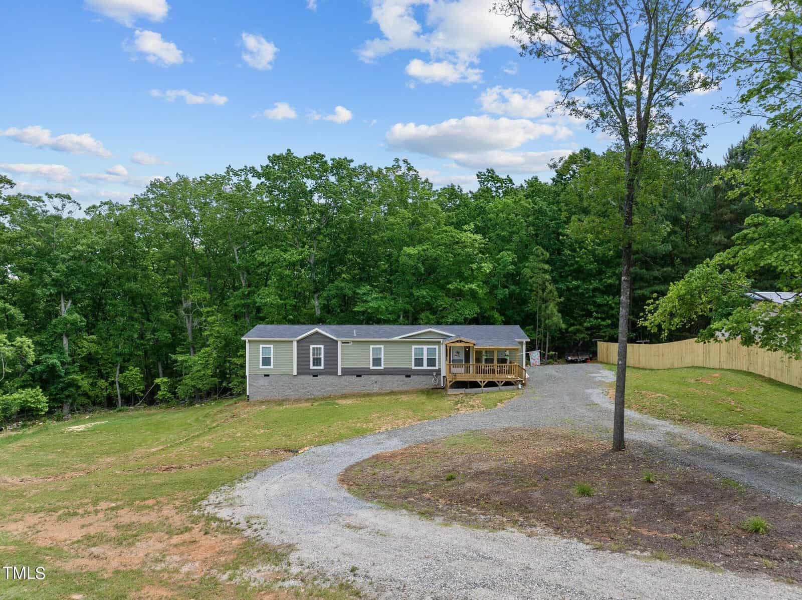 A single-story home with a gray exterior and a screened porch, surrounded by lush trees. A gravel driveway leads to the house, with a wooden fence on the right and a large, grassy yard. The sky is blue with scattered clouds.
