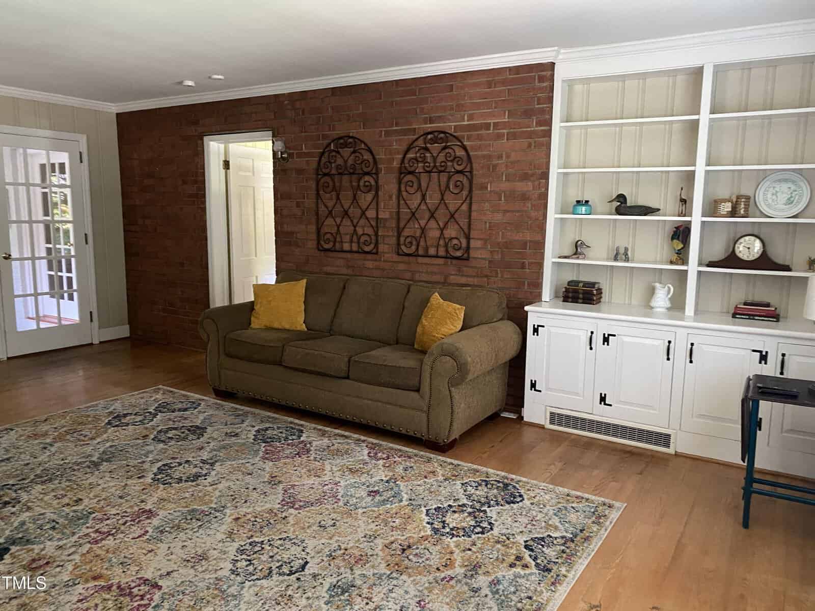 A cozy living room with a brown couch adorned with yellow pillows, positioned against a brick accent wall. Above the couch are two decorative wall hangings. Adjacent is a white built-in bookshelf filled with various items. A patterned area rug covers the wooden floor.