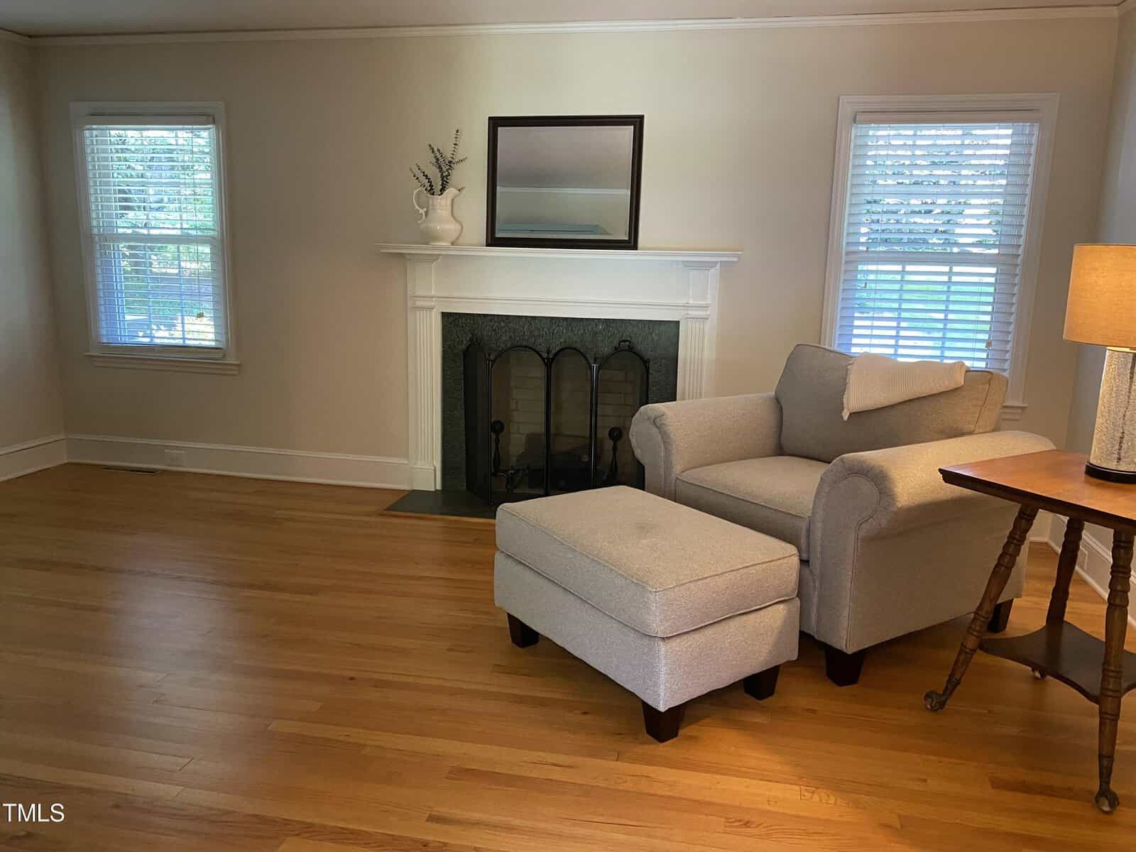 A cozy living room with light wood flooring features a gray armchair and matching ottoman positioned next to a wooden side table. A white fireplace with a black screen and a mirror above it stands between two windows with white blinds. A subtle table lamp adds warmth.