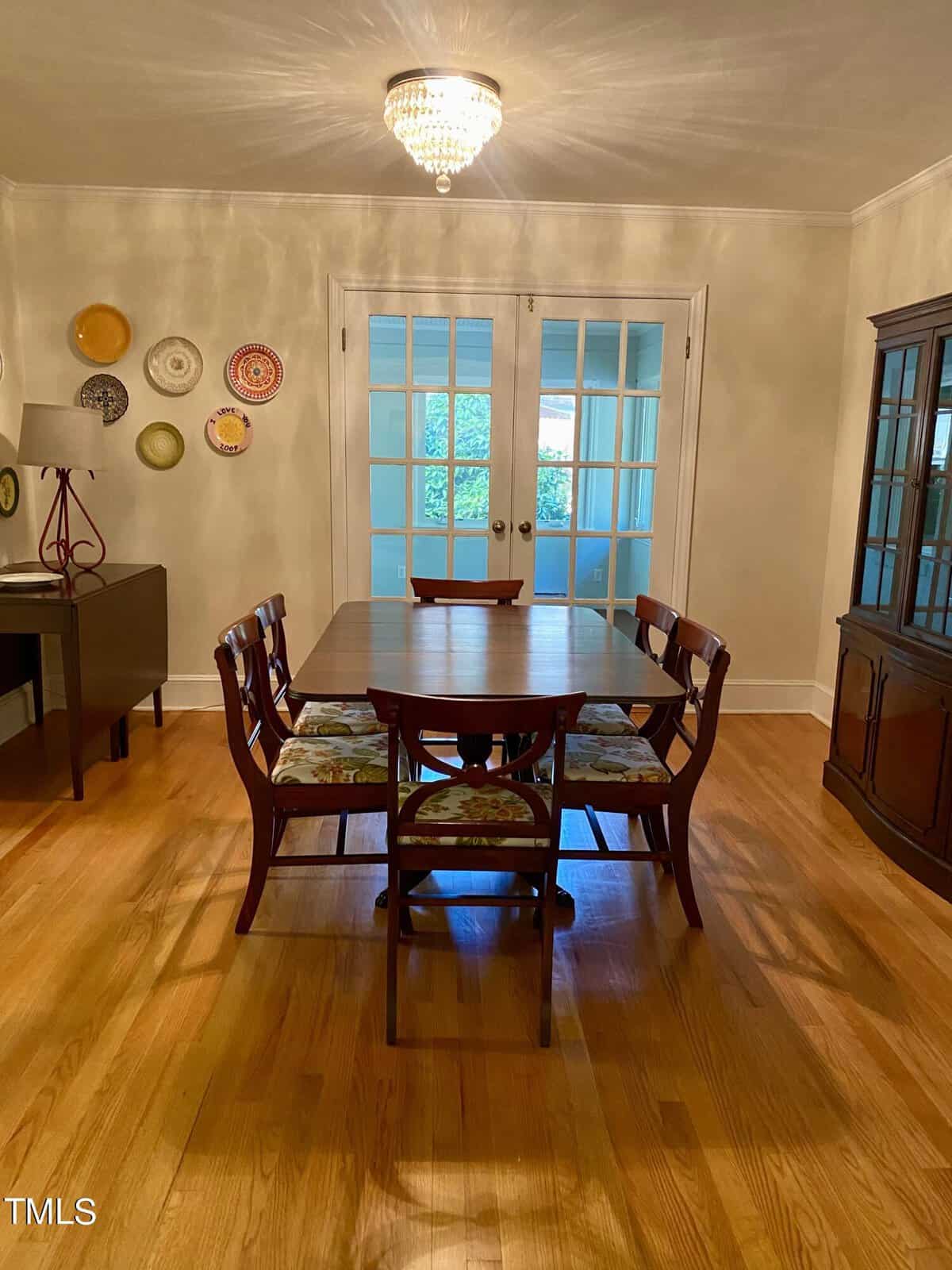 A dining room with a wooden table and six chairs, each with floral-patterned cushions. A chandelier hangs above the table. French doors with glass panes lead to another room. A sideboard with decorative plates on the wall is to the left, and a china cabinet is to the right.
