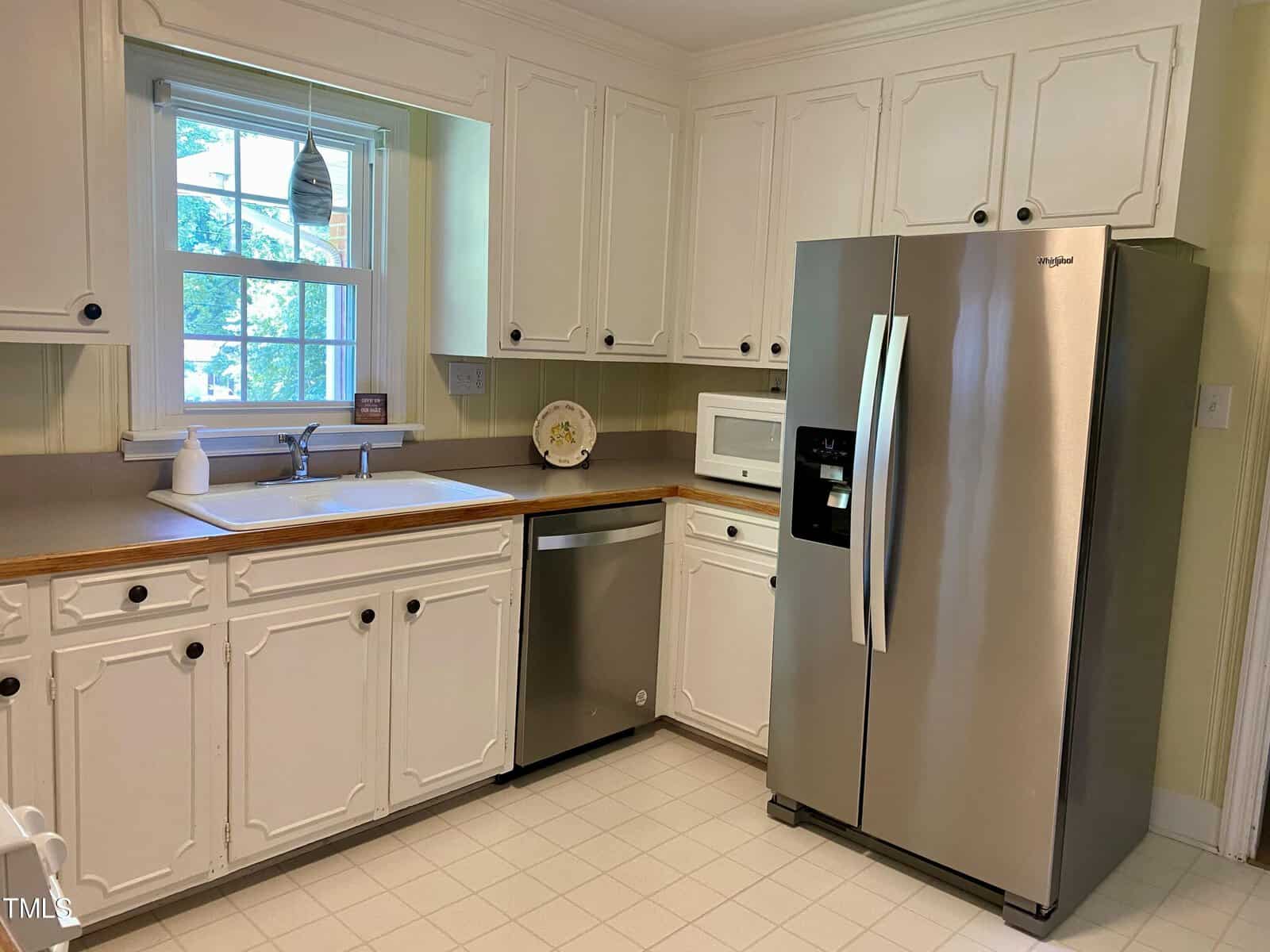A kitchen with white cabinetry, beige countertops, and stainless steel appliances, including a refrigerator and dishwasher. A window above the sink lets in natural light. The kitchen also features a white floor and a microwave on a countertop next to the refrigerator.