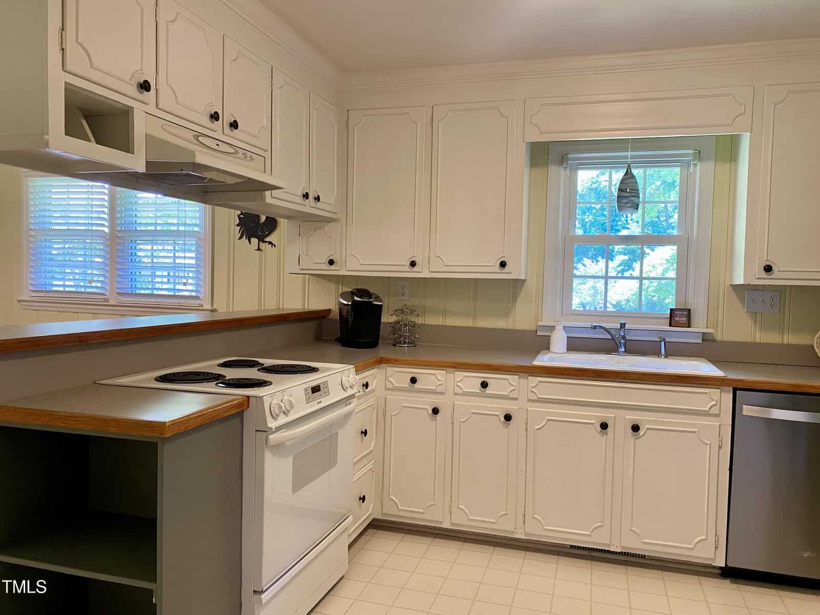 A bright kitchen with white cabinetry and wood-trimmed countertops. It features an electric stove with an overhead vent, a dishwasher, a coffee maker, and a sink beneath a window. The walls are painted a light yellow, and there are two small windows by the stove.