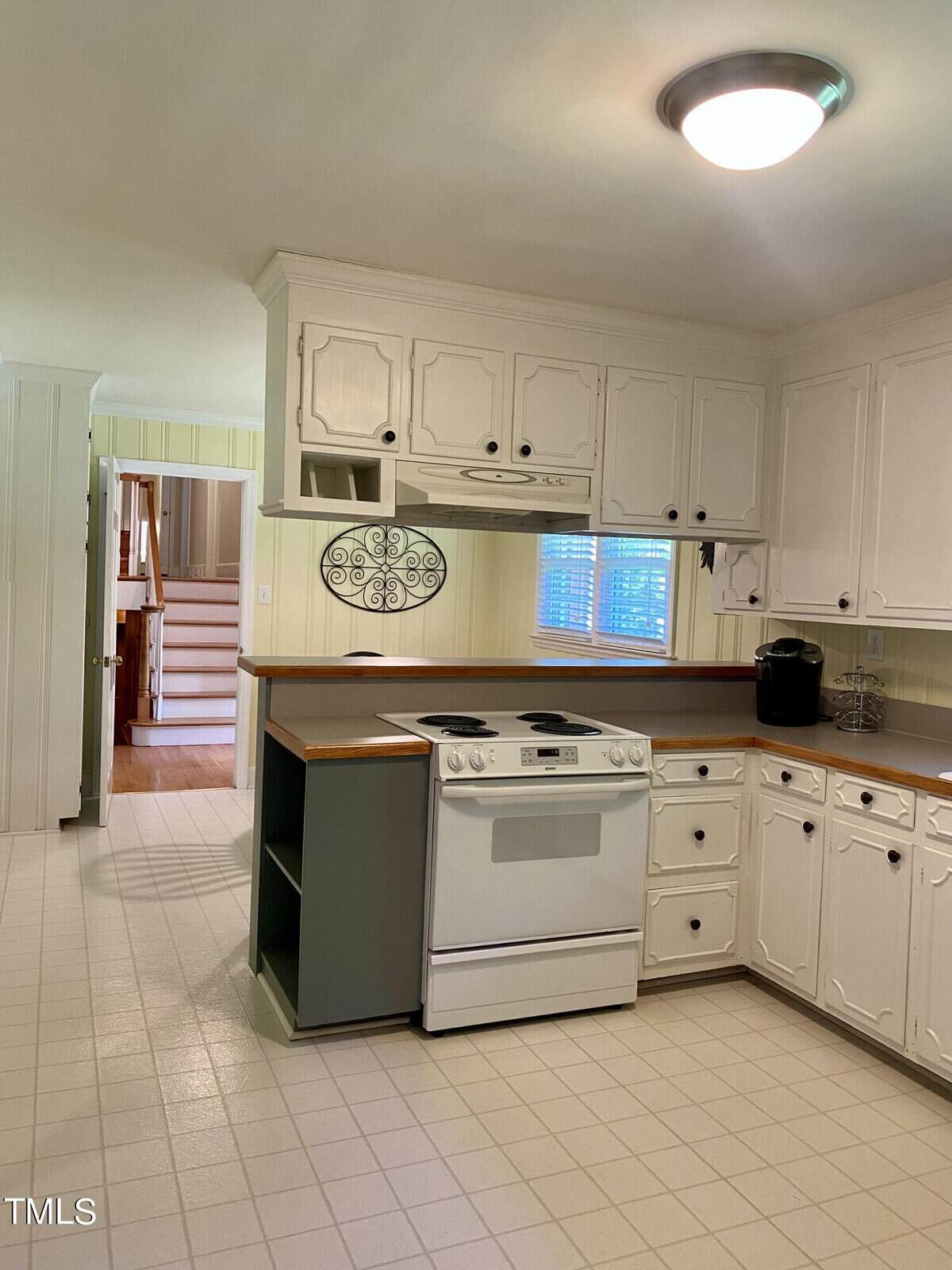 A spacious kitchen with white cabinets, a white oven, and a wooden countertop. The kitchen has a light-colored tiled floor and a circular decorative wall piece. A staircase with wooden steps is visible in the background.