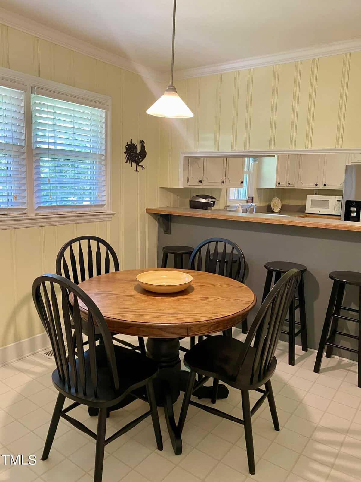 A cozy kitchen area featuring a round wooden dining table with four black chairs. Above the table, a pendant light hangs. In the background, there's a counter with bar stools and a pass-through window to the kitchen, which contains cabinets and appliances.