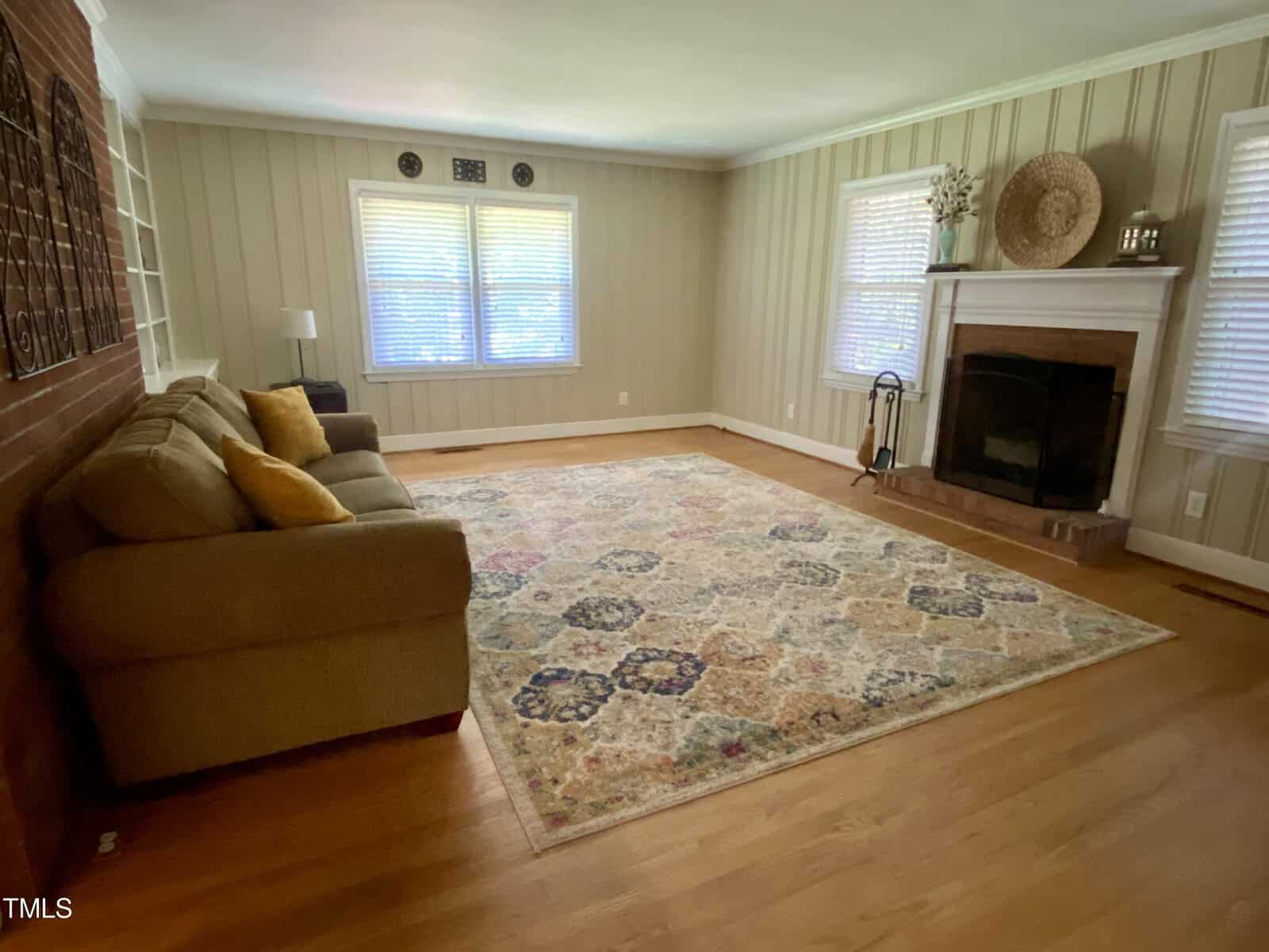 A spacious living room with a beige sofa adorned with yellow pillows, a large patterned rug covering the wooden floor, and a brick fireplace flanked by windows. The neutral-toned walls complement the room's decor, while natural light fills the space through the windows.
