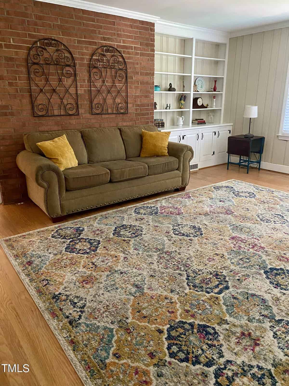 A cozy living room with a green sofa adorned with yellow pillows, set against a brick wall with two decorative metal wall hangings. A colorful patterned rug covers the wooden floor, and built-in bookshelves and a lamp are in the background.