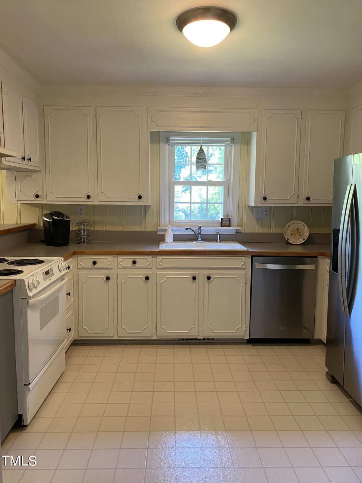 A spacious kitchen with white cabinets, a stainless steel refrigerator, a white stove and oven, a dishwasher, and a sink beneath a window. A coffee maker is on the counter. The floor is covered with light-colored tiles. Warm lighting from a ceiling fixture brightens the room.