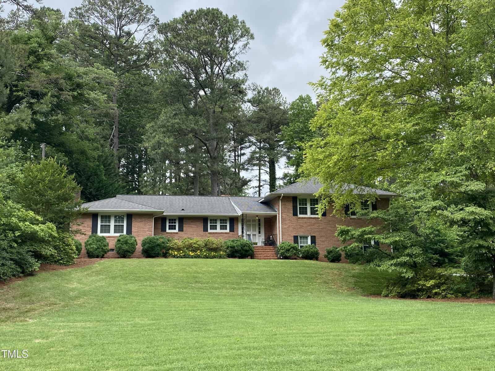 A single-story brick house with a wide front lawn, surrounded by tall trees and shrubs. The house features a central front entrance with a small staircase leading up to it, and has a well-maintained green lawn in the foreground.