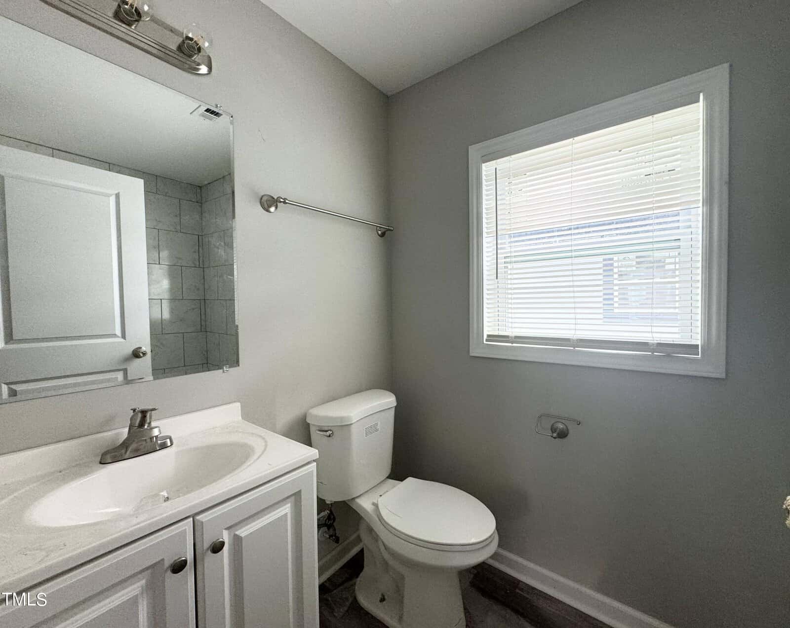 Located at 409 Spring Court, Henderson NC, this small, clean bathroom features light gray walls and a white vanity with a sink and mirror. It includes a white toilet, towel bar, and window with white blinds, allowing light to stream in. The dark wood-patterned flooring complements the space beautifully.