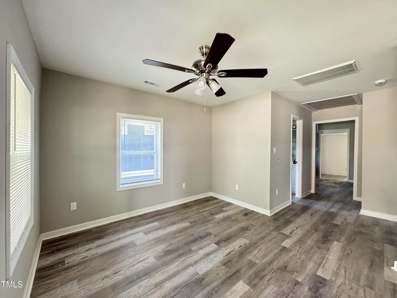 A bright, empty room with light gray walls, large windows, and dark wood flooring. A ceiling fan with lights is centered in the room. A hallway and additional rooms can be seen in the background.