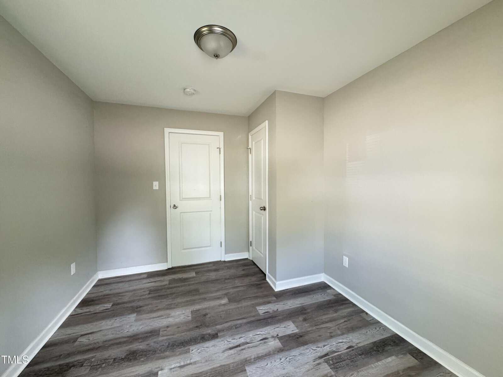 A small, empty room with light gray walls and dark gray hardwood flooring. The room features a flush mount ceiling light fixture, a closed white door, and a closed white closet door. The room appears clean and bright with ample natural light.