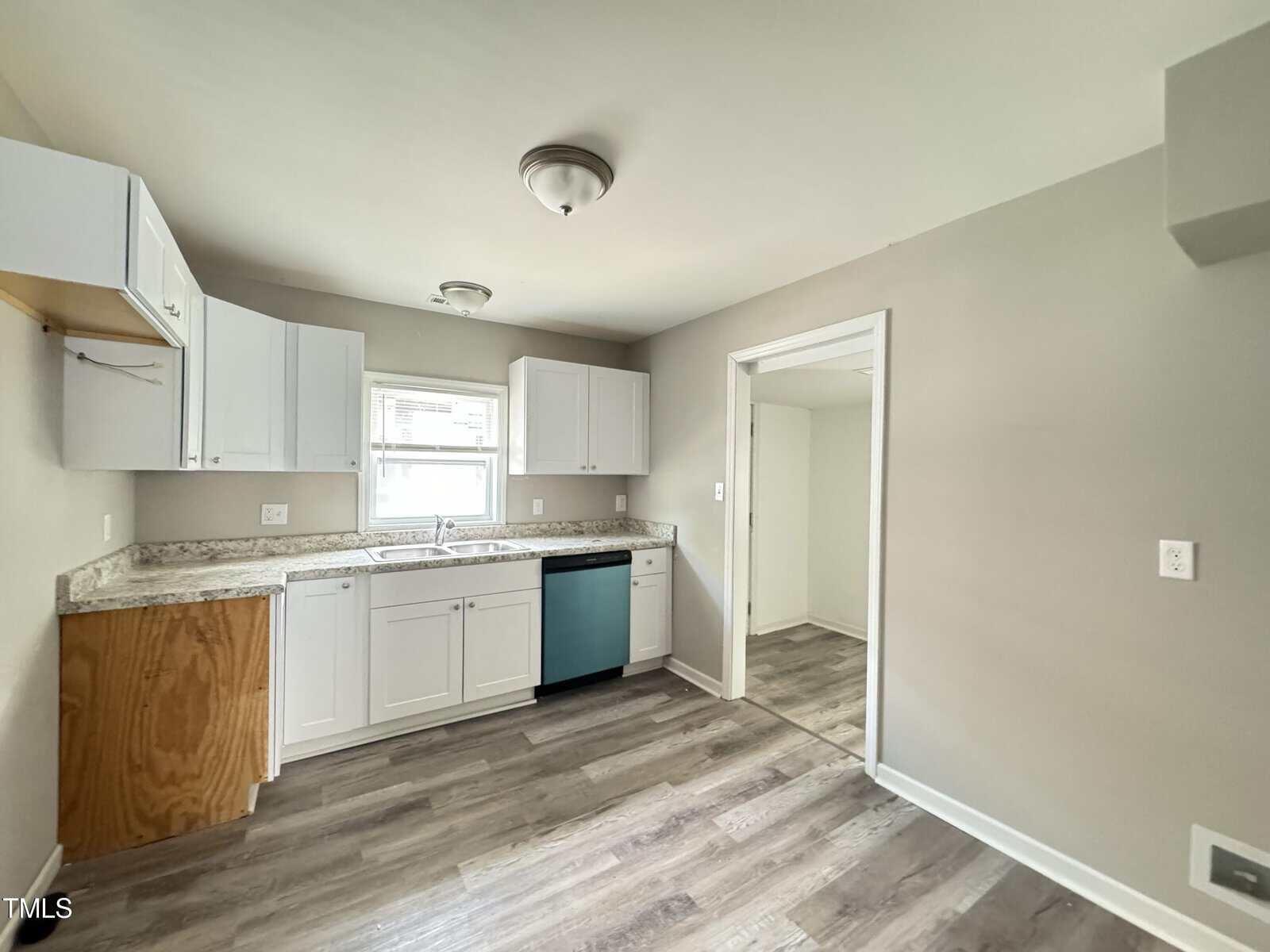 A modern kitchen with light gray walls and wood-look flooring. It features white cabinets, a stainless steel sink under a window, and a white ceiling light fixture. There is a pass-through opening to an adjacent room. One cabinet door is missing.