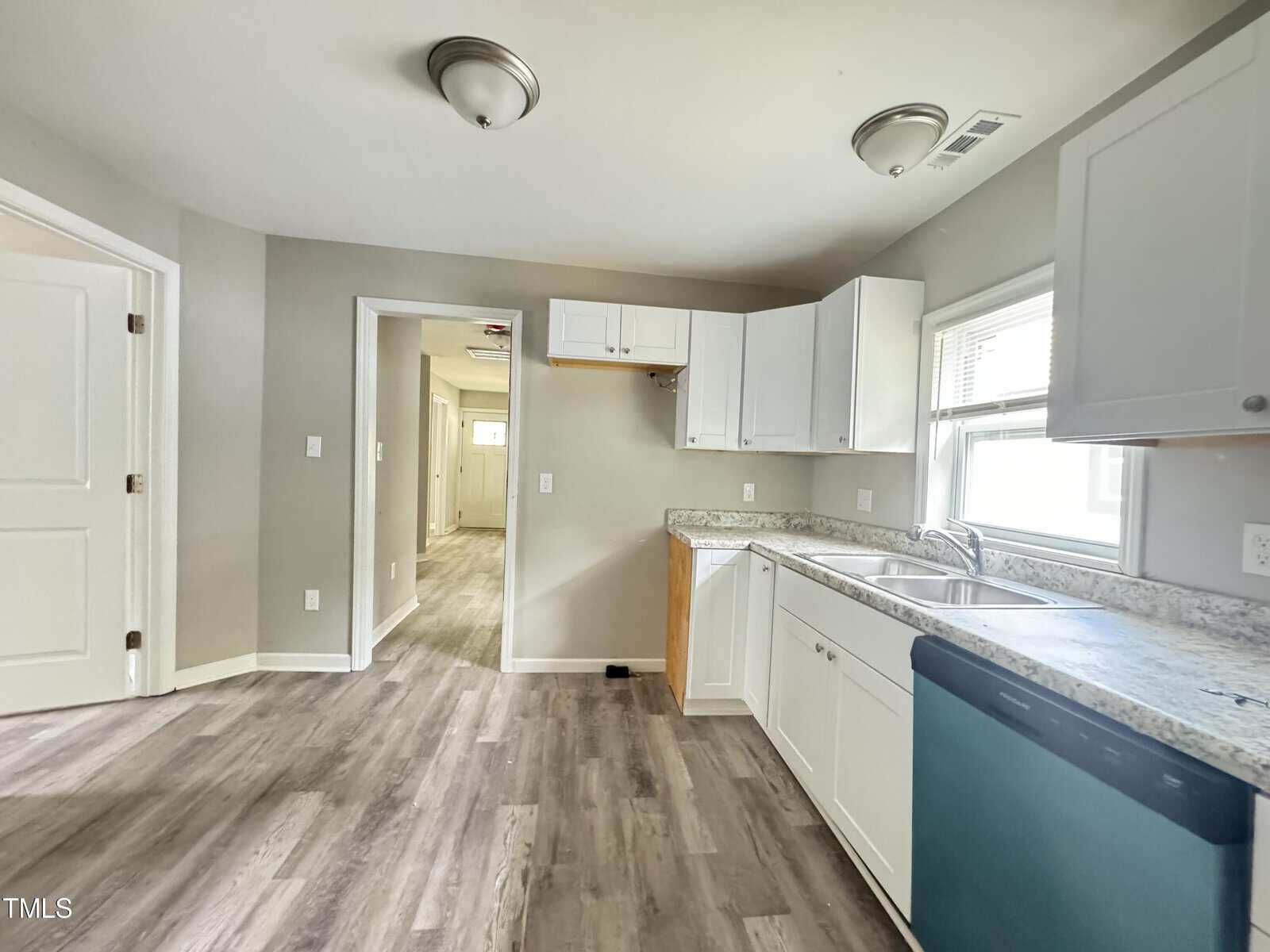 A modern kitchen with white cabinets, light gray walls, and light wood flooring. There is a double stainless steel sink, a dishwasher, and a large window above the sink. A hallway entrance is visible on the left, leading to another room.