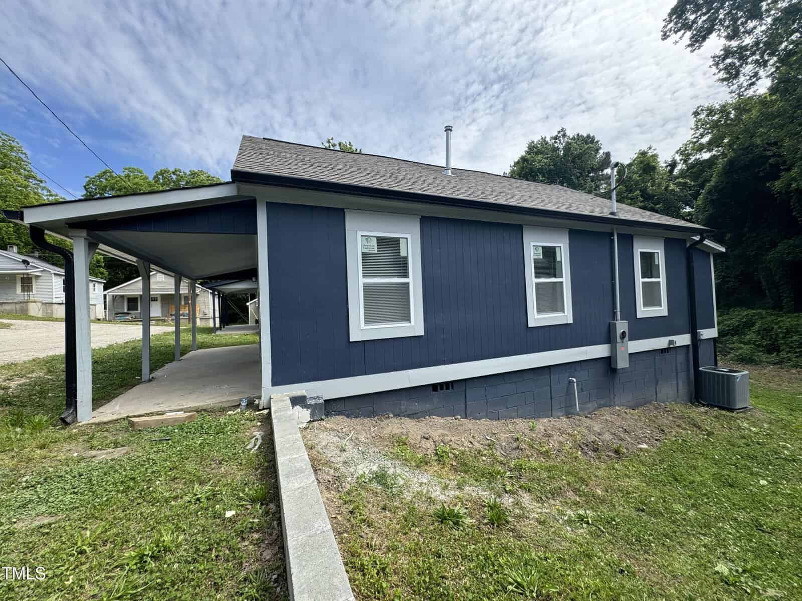 A small, single-story house with dark blue siding and white trim is pictured. The house features a carport on the left side, and there are two visible windows on the side facing the camera. The roof is gray, and there is a small lawn with some greenery.