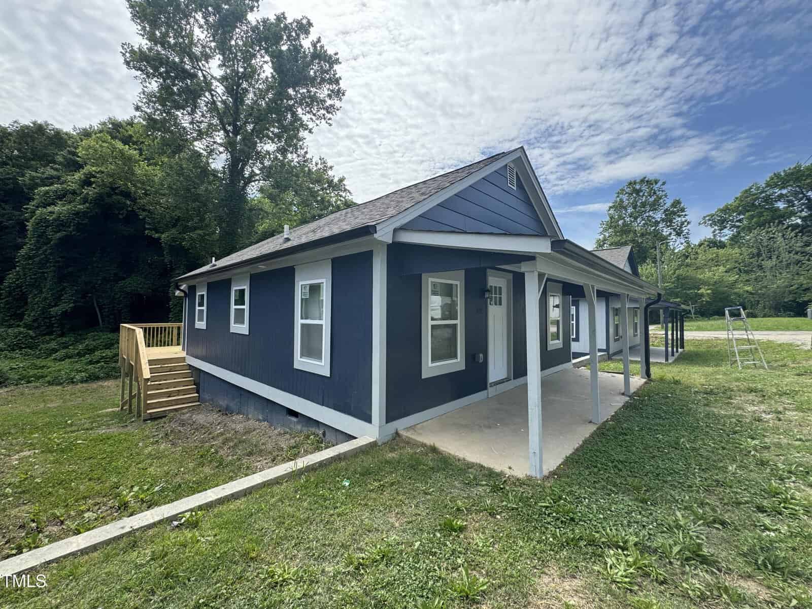 A small blue house with white trim, featuring a covered front porch and a wooden deck at the side. The house is surrounded by a grass lawn and trees, and a partly cloudy sky is visible above. A metal ladder and a ditch are seen in the yard.