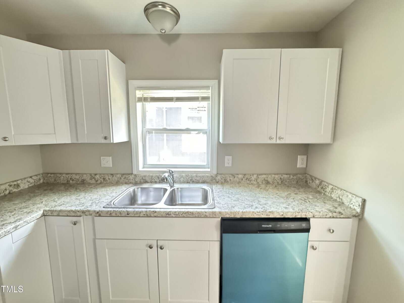 A kitchen features white cabinets, marble countertops, a double stainless steel sink under a window with blinds, and a stainless steel dishwasher with a blue protective cover. The walls are painted in a light neutral color.