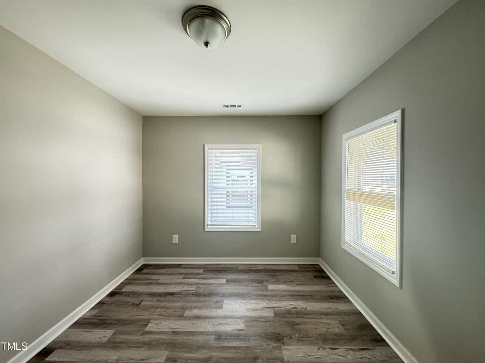 A small, empty room with light gray walls and wooden flooring. The room has two windows with white blinds, allowing natural light to enter. A ceiling light fixture is centered above. The overall space is clean and minimalistic.