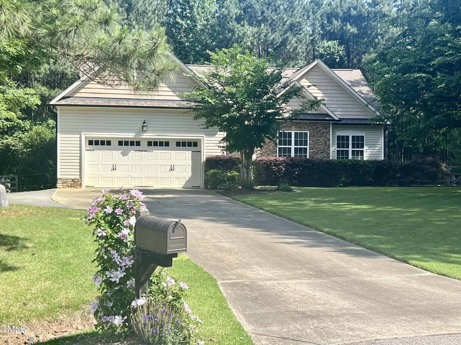 Nestled in Franklinton, this suburban house at 1203 Bayview Court boasts a beige exterior, attached garage, and sloped driveway. Lush trees and shrubs grace the yard, while a mailbox and blooming pink flowers adorn the entrance to the driveway.