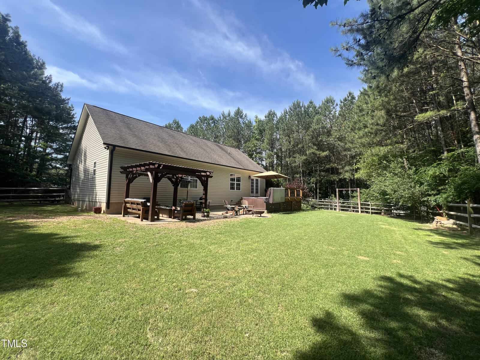 A single-story house with a gray roof sits in a spacious backyard surrounded by tall trees. A wooden pergola covers patio seating, and a wooden deck with a hot tub is nearby. The sky is clear and blue, casting shadows on the manicured lawn.