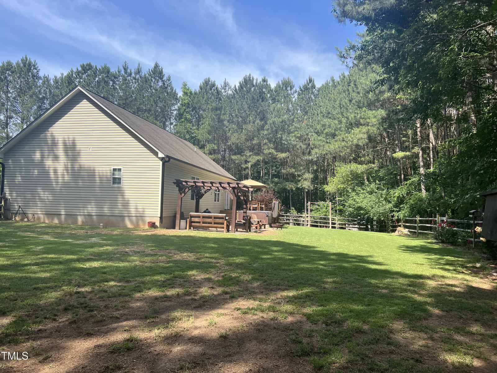A backyard with a grassy area, a modern house on the left, and a wooden pergola with outdoor seating. Trees line the background under a clear blue sky, providing a peaceful, natural setting.