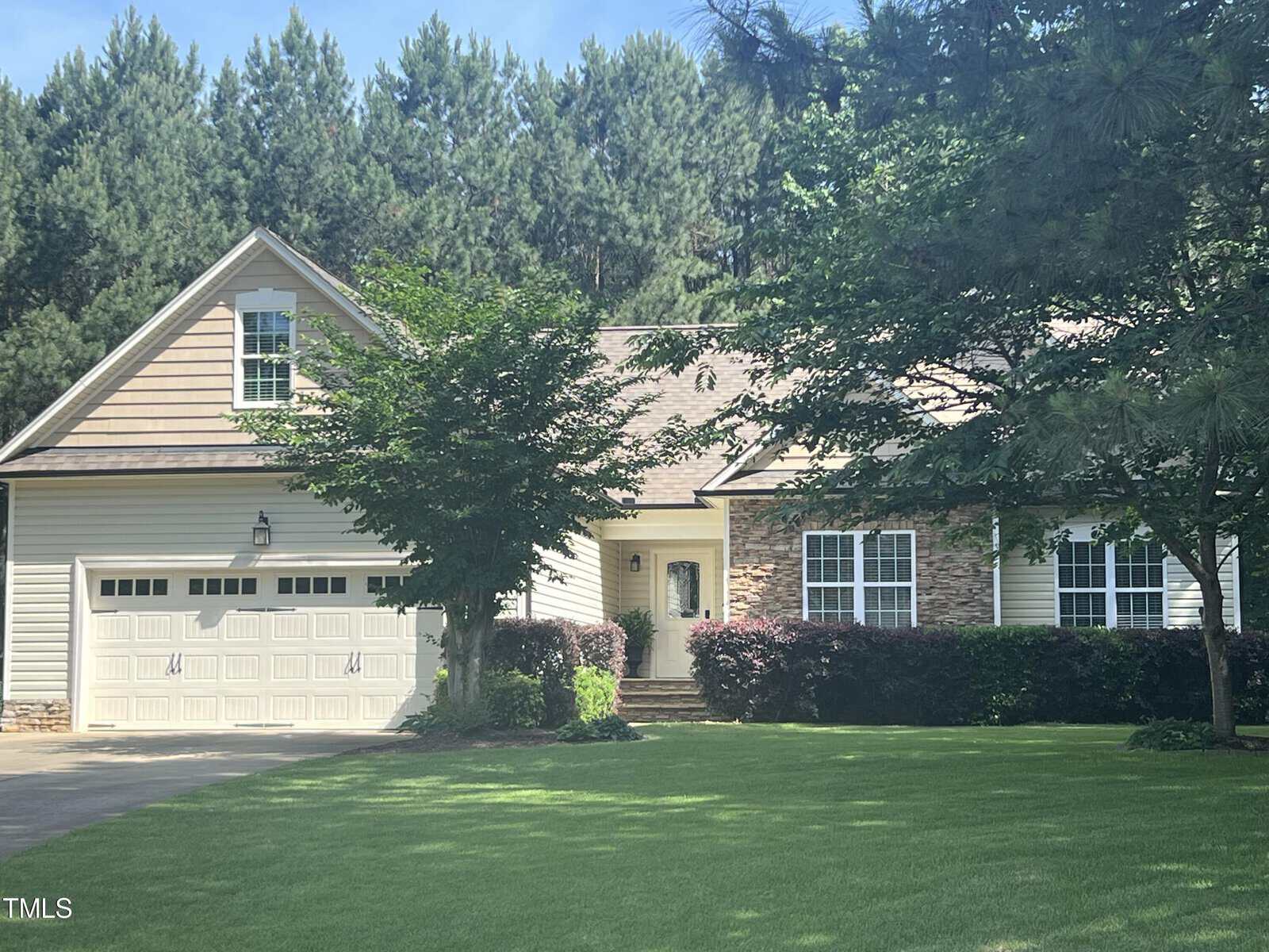 A suburban house with a two-car garage and light-colored siding sits among trees. There's a well-kept lawn in front, and a stone pathway leads to the entrance. Tall trees form a lush, green backdrop.