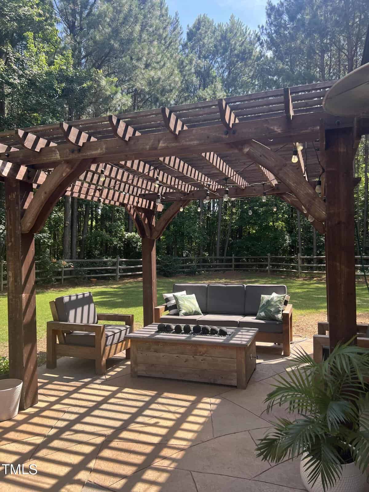 Wooden pergola with seating area underneath, featuring a gray sofa with green pillows, wooden chairs, and a central rectangular table. Sunlight creates shadows on the patio floor. Lush trees and a wooden fence provide a serene background.