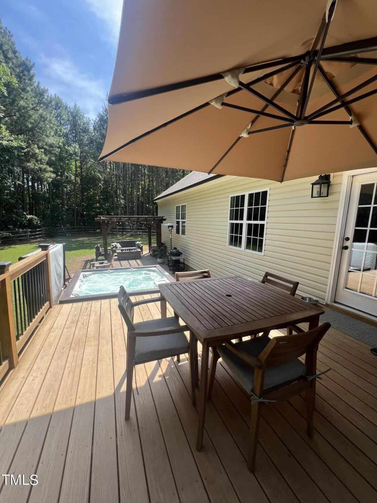 A wooden deck with a patio table and chairs under a large umbrella. Adjacent to the table is a hot tub. The house is light yellow with white-framed windows, and in the background, there is a lush green forest.