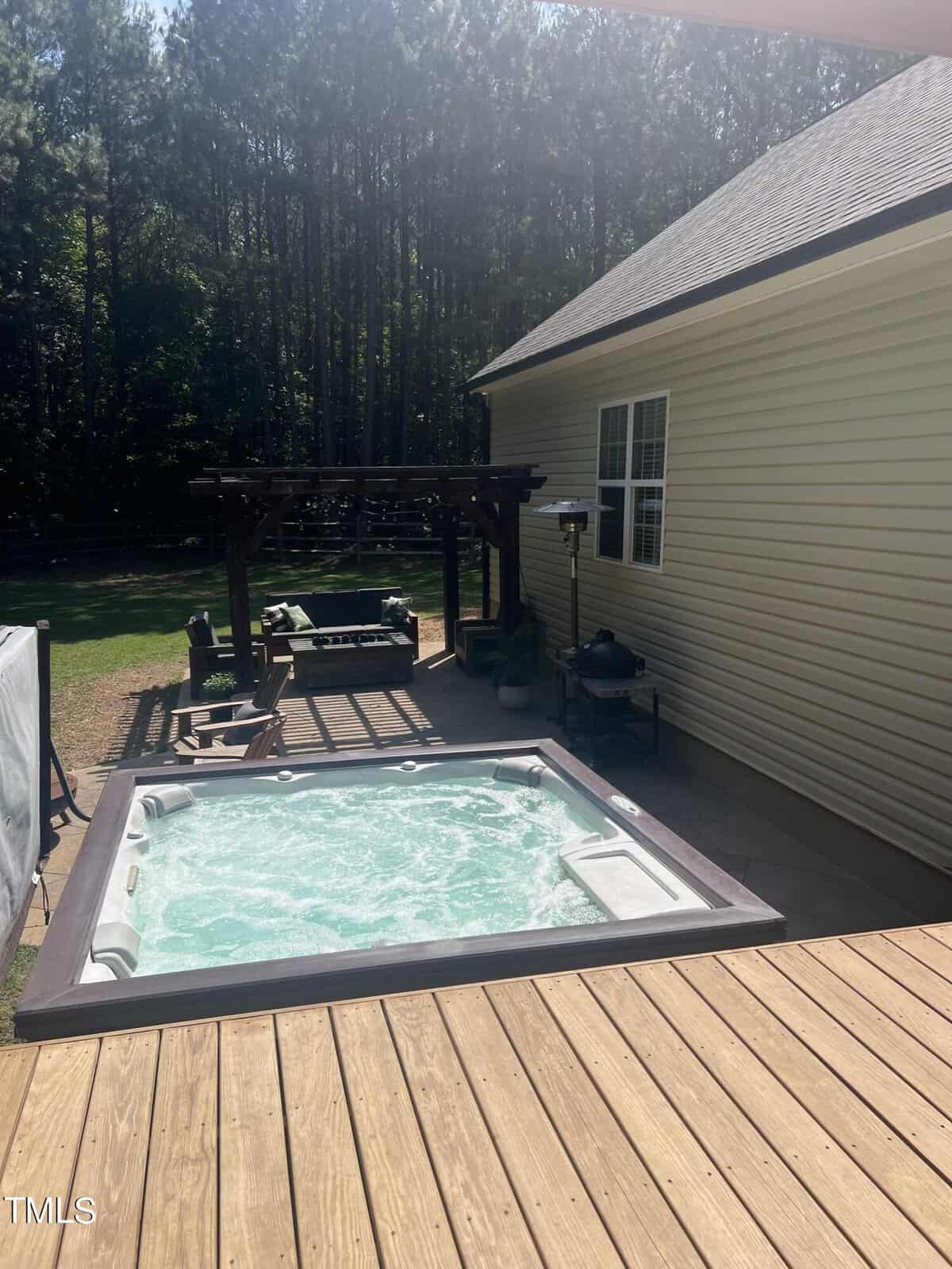 A wooden deck with an in-ground hot tub filled with bubbling water. In the background, there's a seating area with chairs and a small table under a pergola. A yellow house and a line of tall trees are visible beyond the deck.